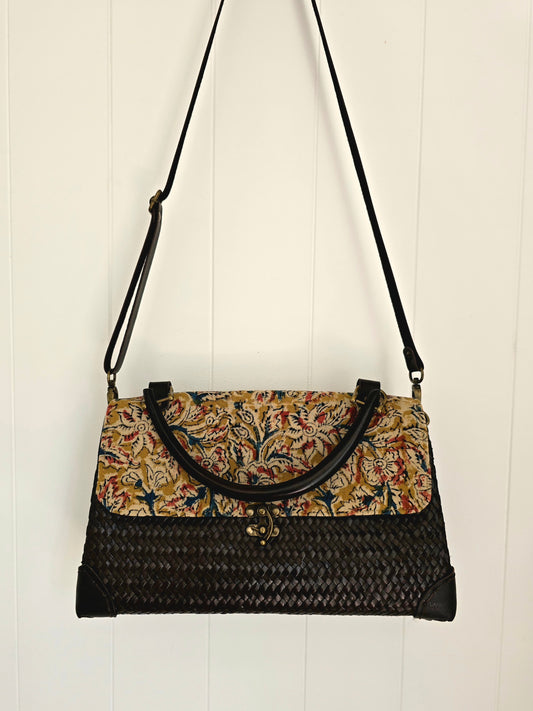 A black woven handbag with a floral patterned top flap and black leather trim, hanging against a white paneled wall.