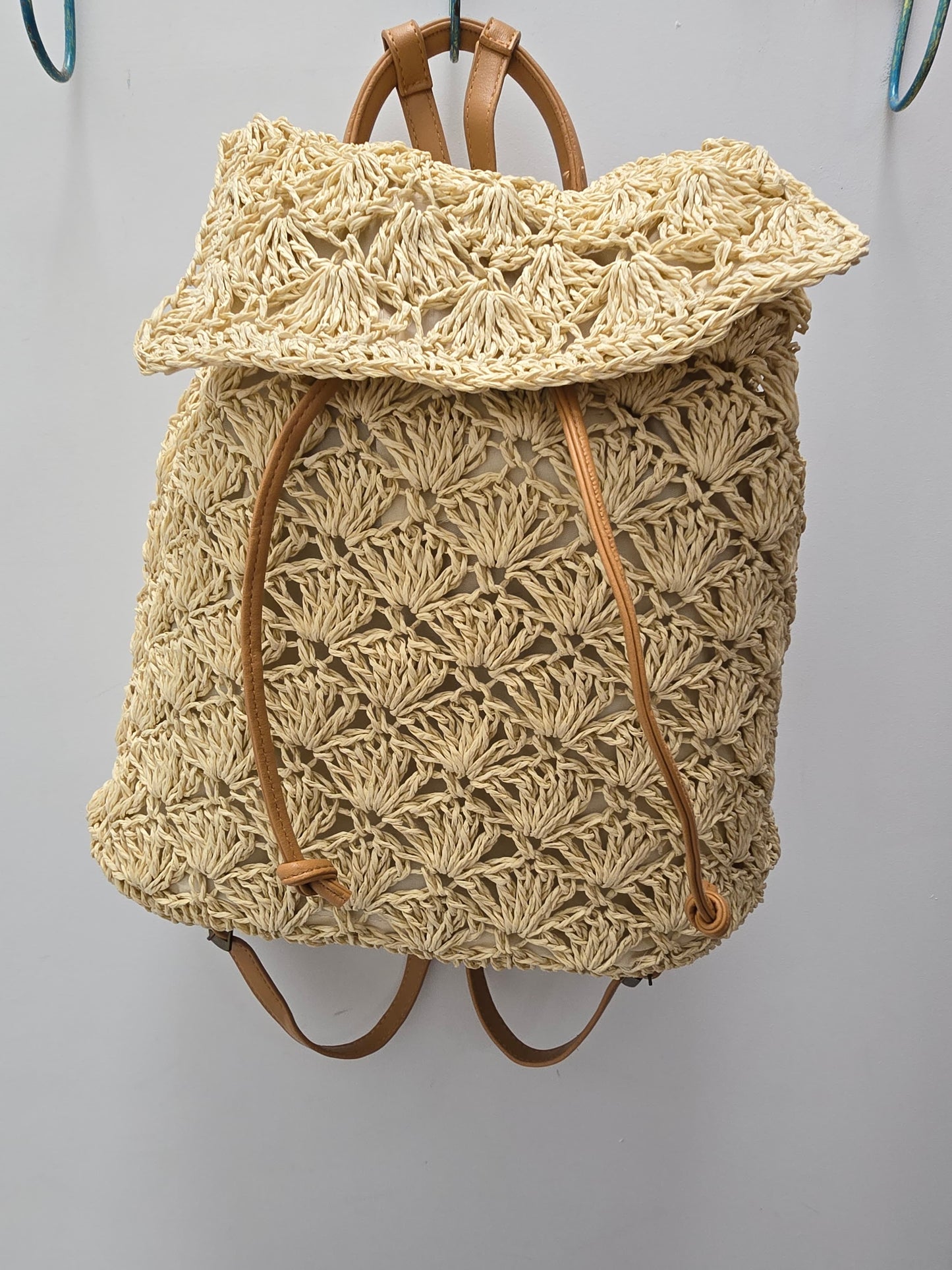 A natural straw woven backpack with a textured, fan-like pattern and tan leather straps, hanging against a plain light gray wall.
