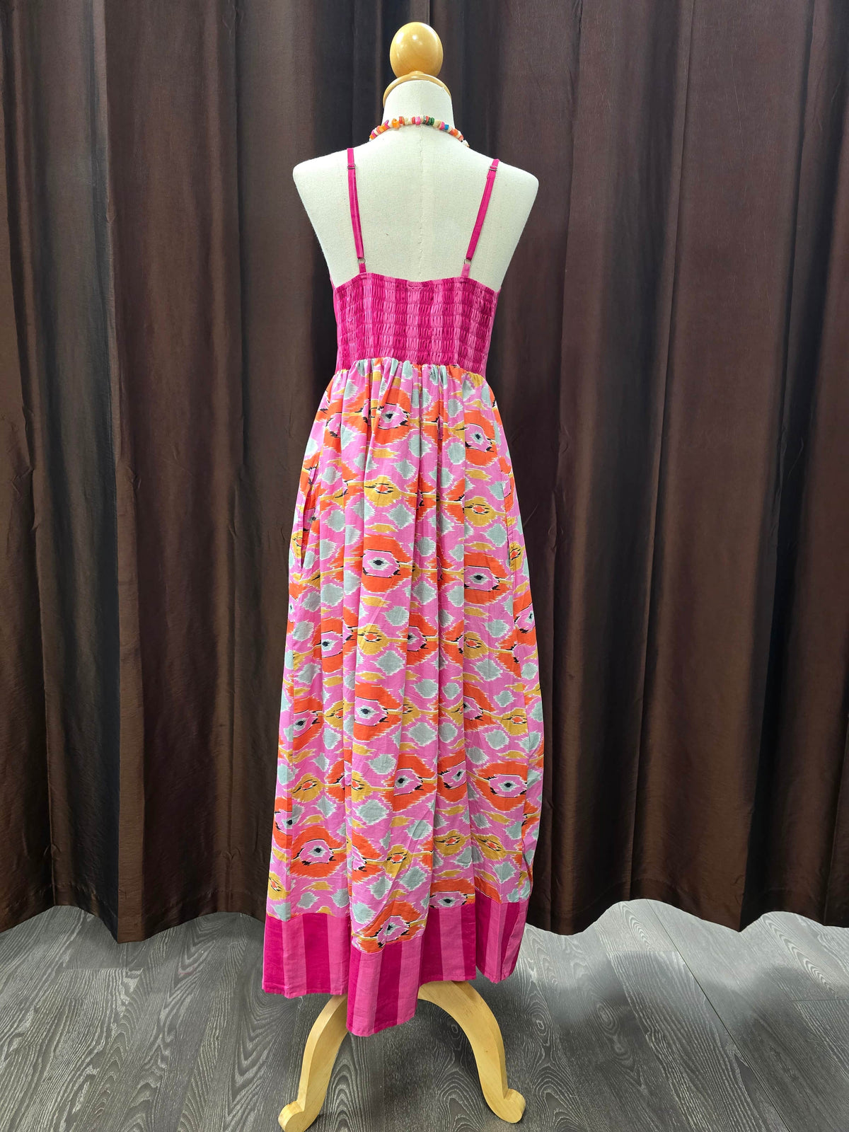 A sleeveless maxi dress displayed on a cream dress form mannequin, featuring a bright pink pleated bodice and a lower skirt with a colorful cartoon fish pattern on a light background, set against a dark brown curtain backdrop.