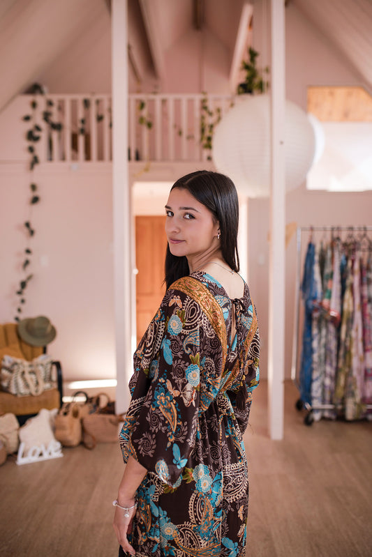 A woman with long black hair stands in a boutique interior wearing a brown patterned kaftan dress with blue, gold, and beige motifs. The background features a wooden loft staircase, warm lighting, and clothing racks.