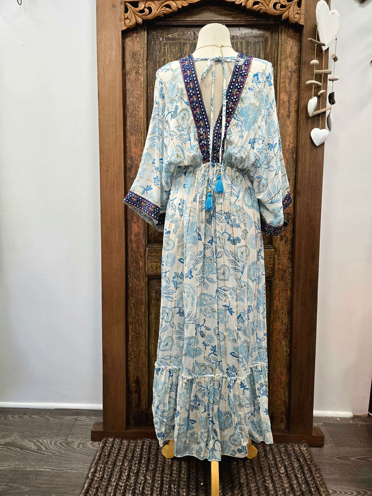 A long kaftan-style dress displayed on a mannequin in front of a carved wooden door. The dress has a light blue and white floral print with dark blue and purple beaded trim along the V-neckline and sleeve cuffs, and blue tassels at the waist.