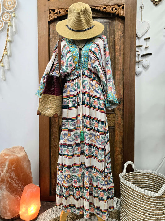 A bohemian-style maxi dress with a colorful paisley and striped pattern, styled on a mannequin with a straw hat and shoulder bag, set against a rustic wooden door background.