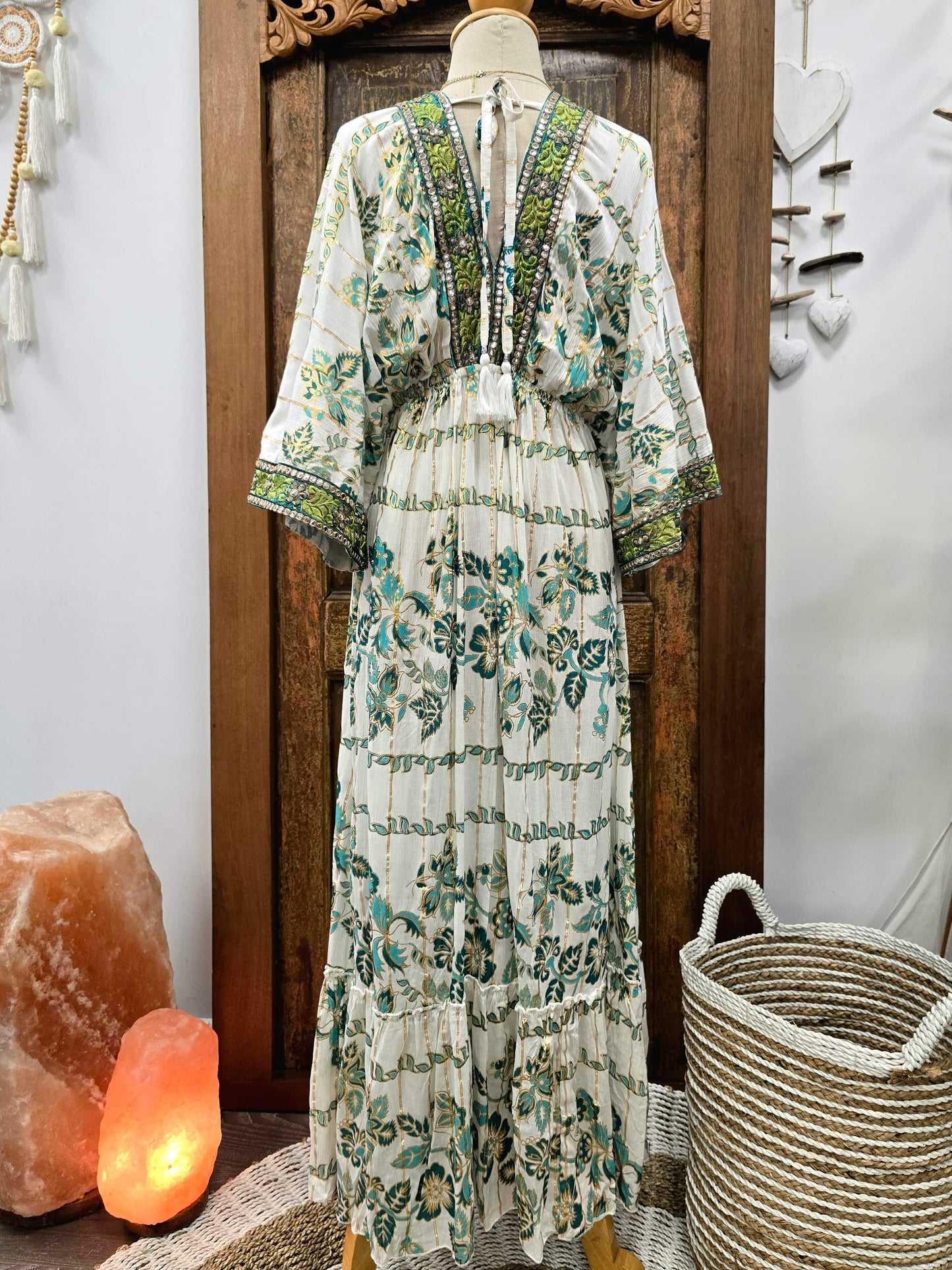 A bohemian-style tiered maxi dress with a green and teal floral print on an off-white base, displayed on a mannequin in front of a dark wooden door. The dress features a V-neckline with green embroidered detailing and wide three-quarter sleeves.