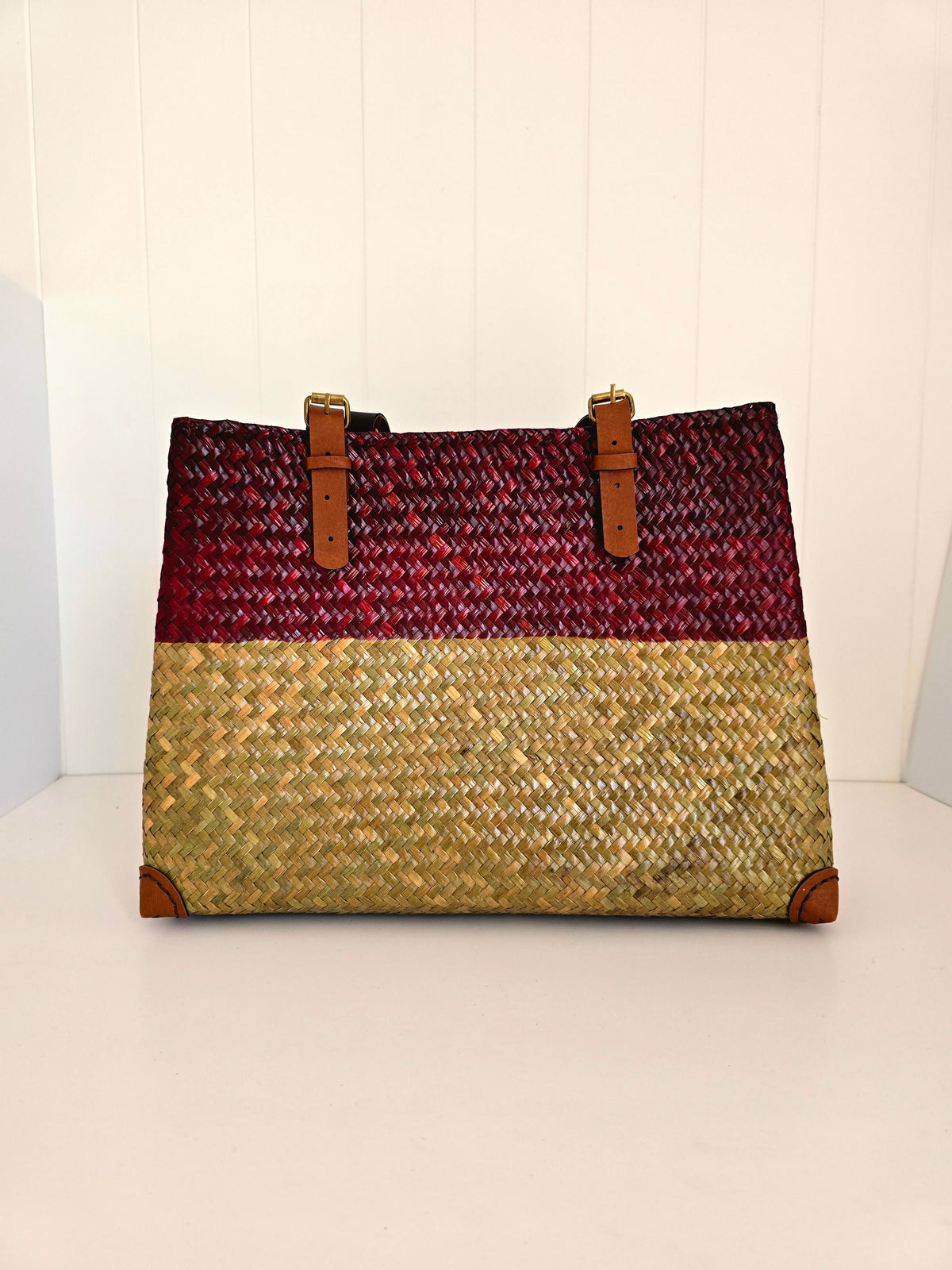 A woven tote bag with a two-tone design, featuring a deep red upper section and a natural beige straw lower section, accented with brown faux leather handles and corner protectors.