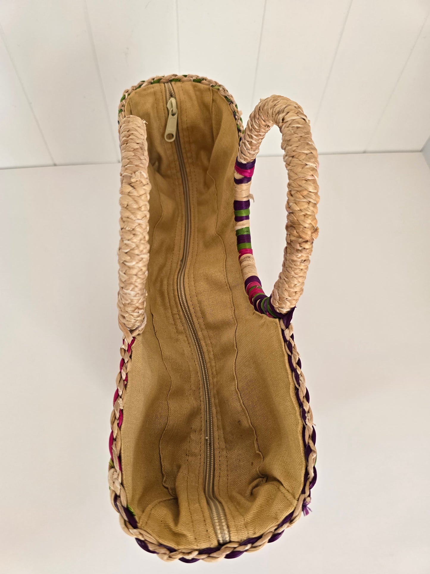 A top-down view of a tan canvas zippered bag with thick braided straw handles and colorful woven trim along the edges, set against a white paneled background.