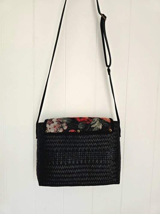 A black woven straw shoulder bag with a floral-patterned tapestry fabric lining on the top flap, hanging against a white vertical wood-paneled wall.
