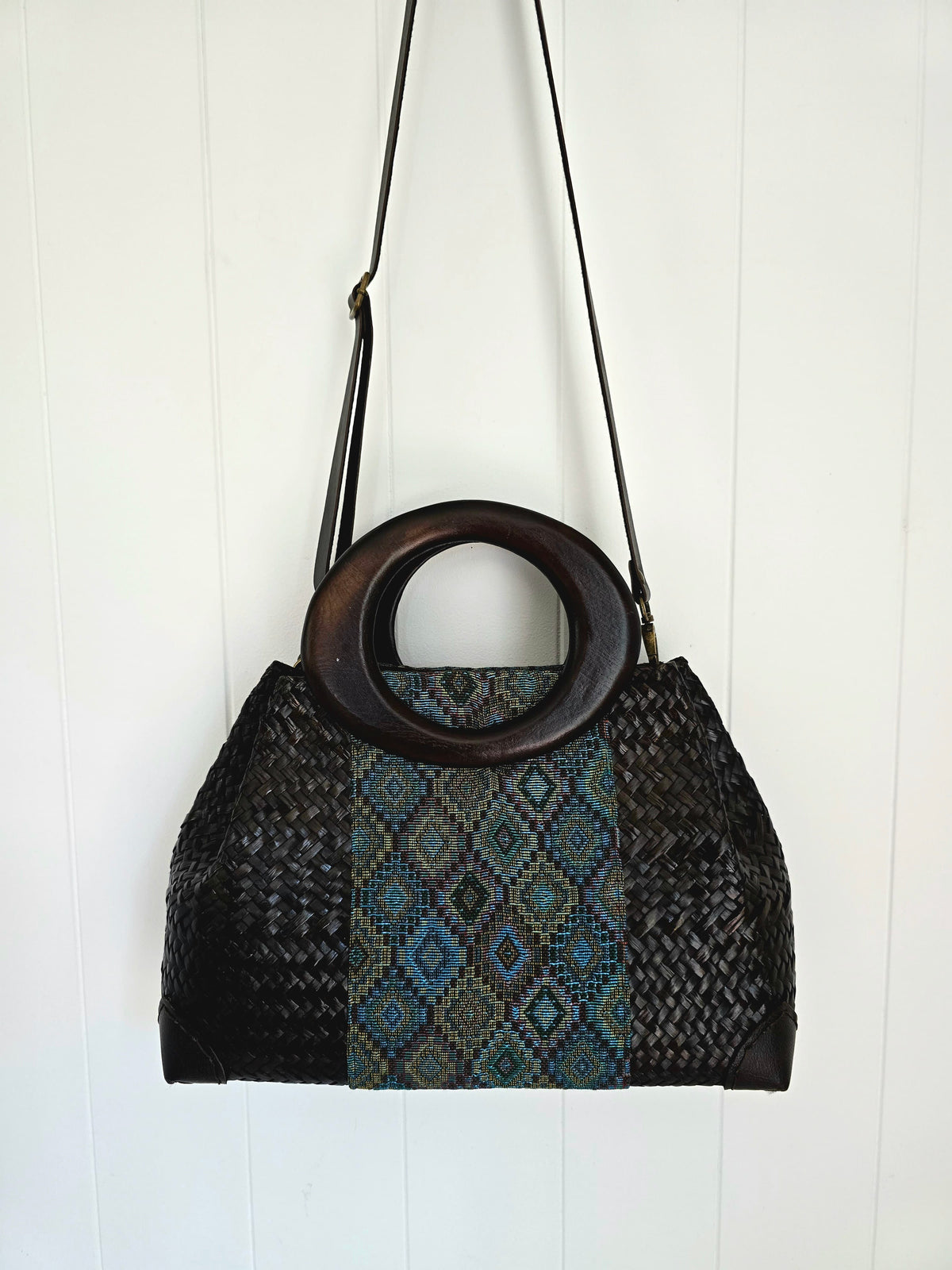 A woven shoulder bag with a circular wooden handle, featuring a black braided body and a central panel with a blue, green, and gold geometric diamond pattern, hanging against a white paneled wall.