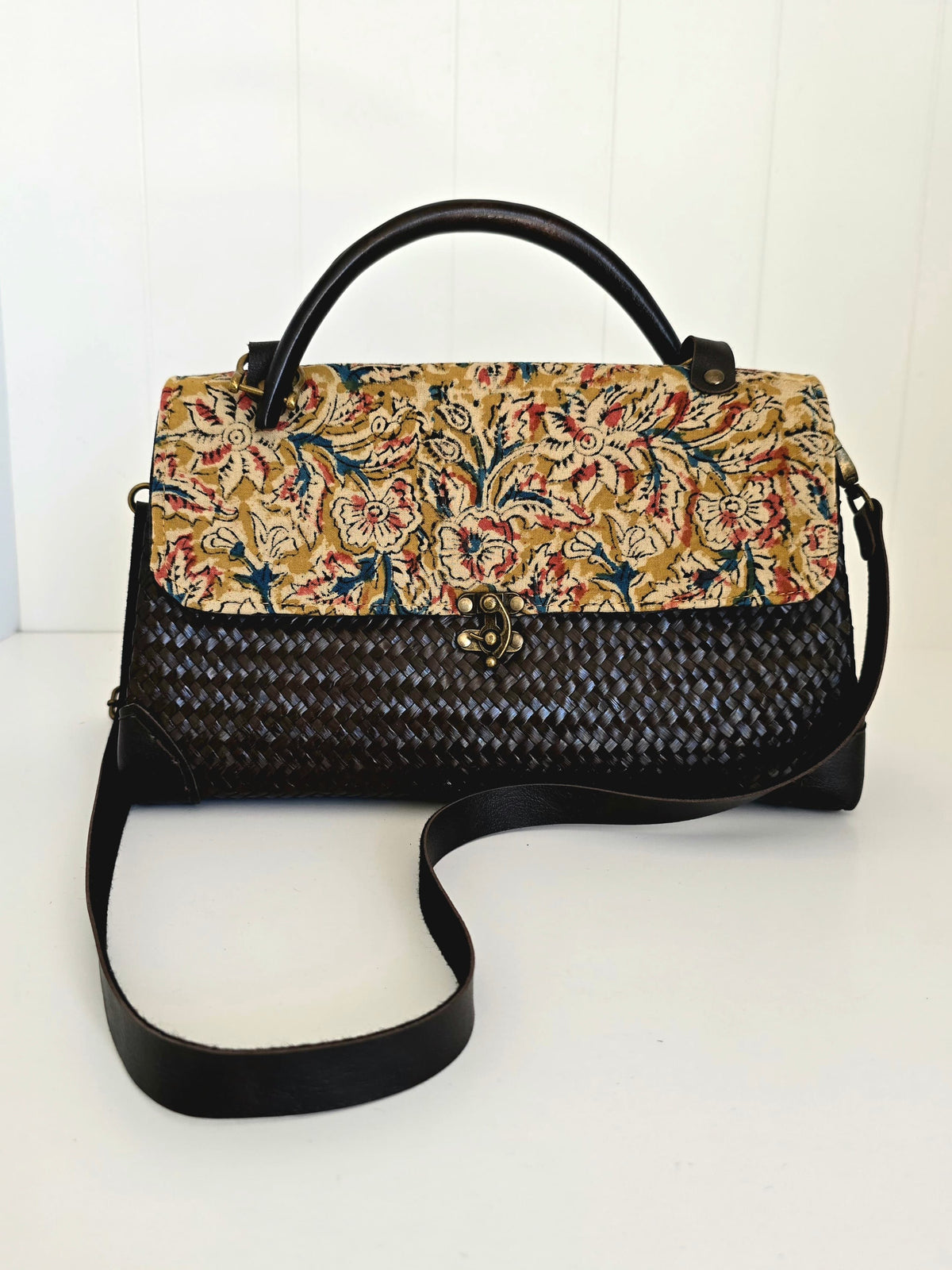 A structured handbag with a black woven body, black leather top handle and shoulder strap, and a floral tapestry-style flap in beige, red, blue, and green.
