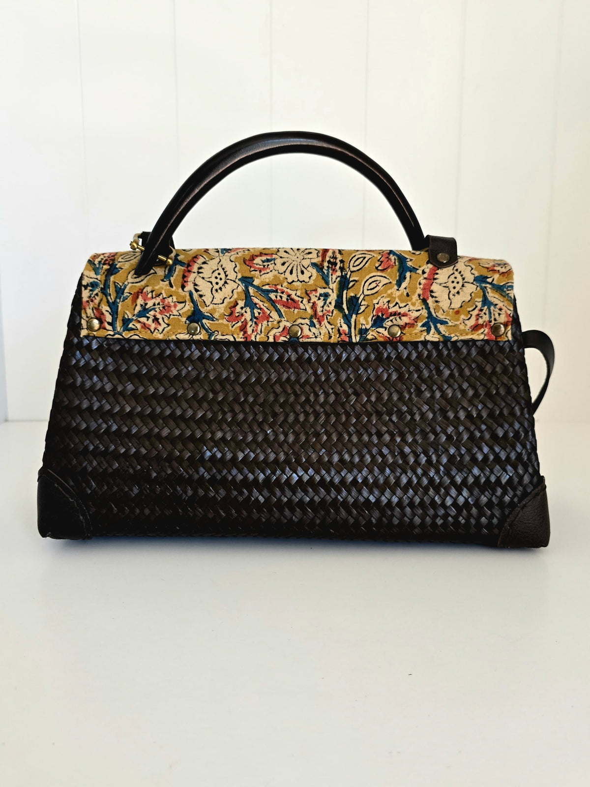A structured handbag with a black woven body, black top handle, and a floral patterned flap in beige, red, and blue, set against a white paneled background.