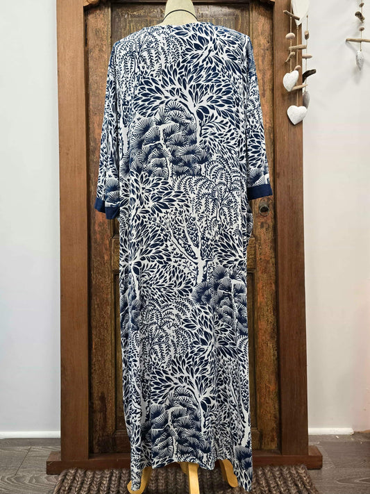 A long-sleeved maxi dress with a blue and white tree branch pattern, displayed on a mannequin in front of a rustic wooden door.