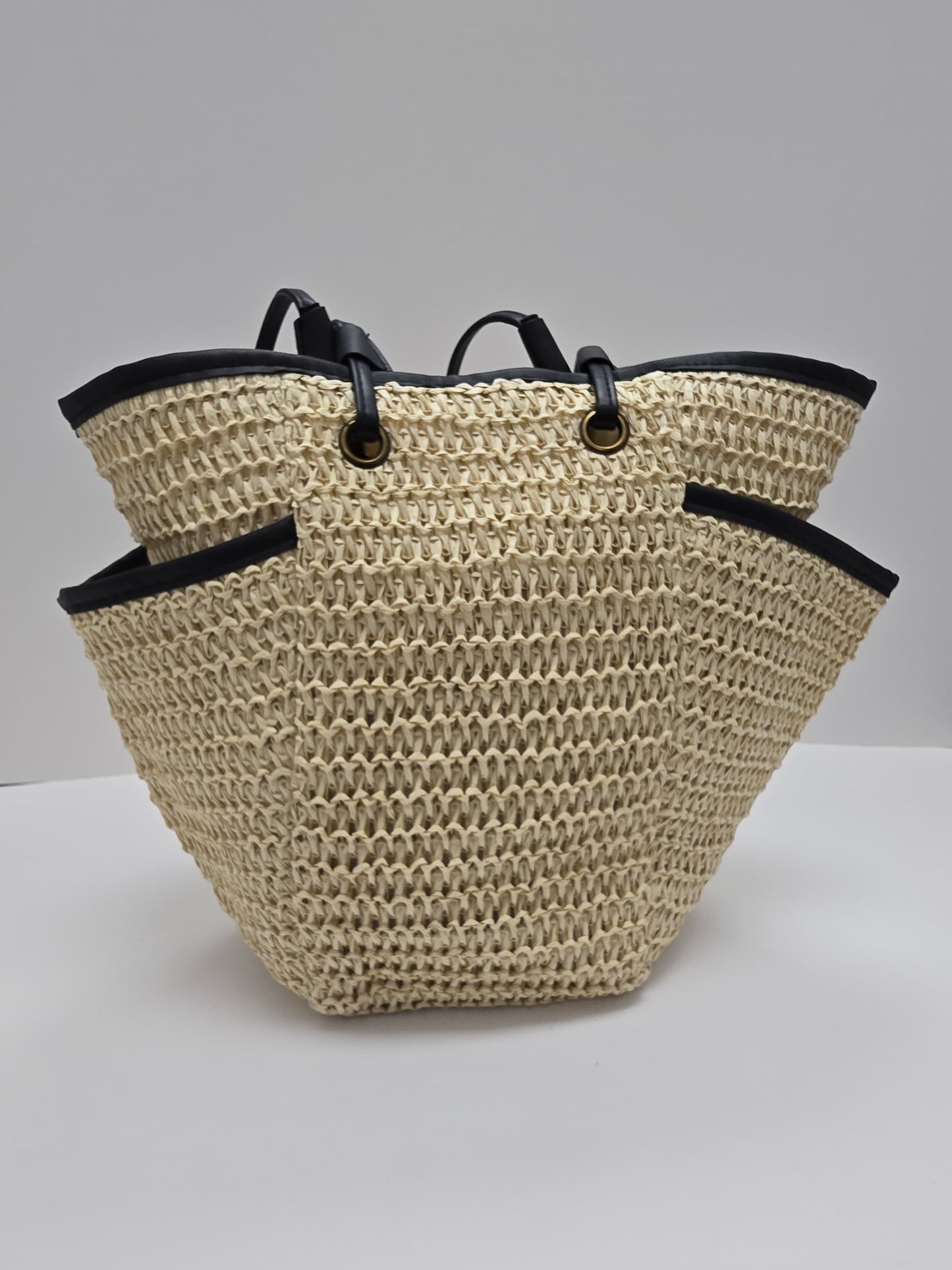 A woven straw tote bag with black trim and double shoulder straps, photographed against a neutral gray studio background.