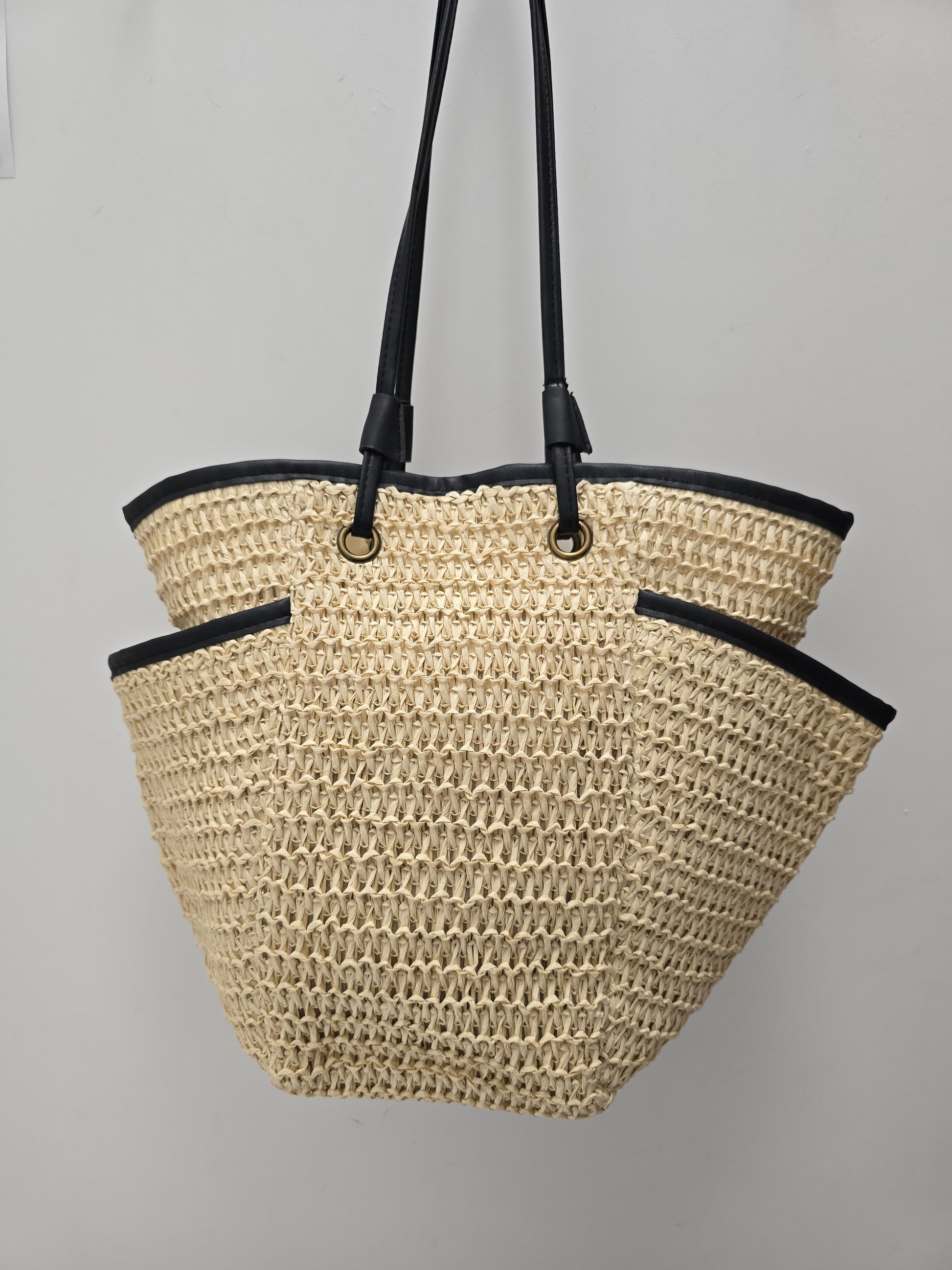 A woven straw tote bag with black trim and black shoulder straps, hanging against a plain light gray wall.