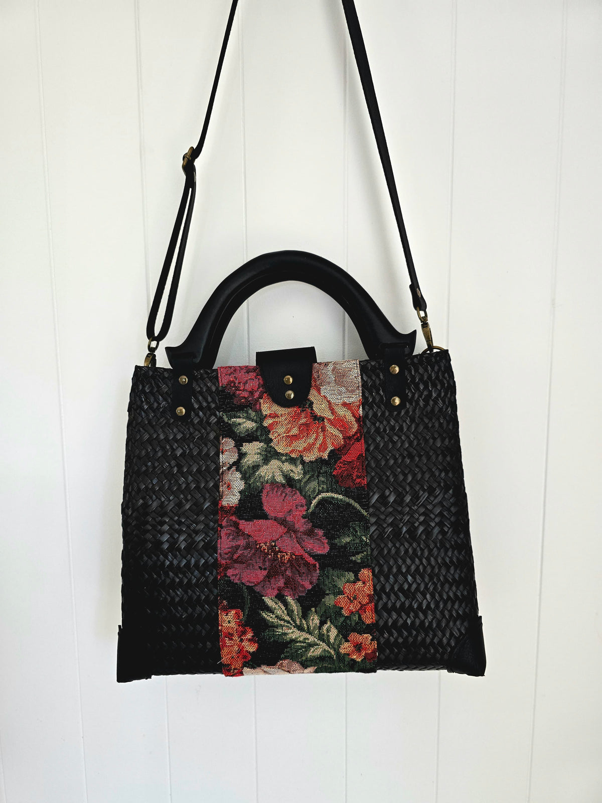 A black woven handbag with a floral tapestry panel in the center, featuring red, pink, and orange flowers with green leaves, and a black top handle and shoulder strap, shown hanging against a white paneled wall.