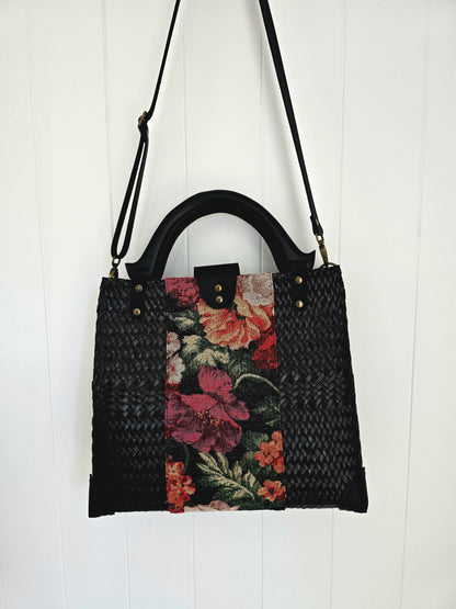 A black woven handbag with a floral tapestry panel in the center, featuring red, pink, and orange flowers with green leaves, and a black top handle and shoulder strap, shown hanging against a white paneled wall.