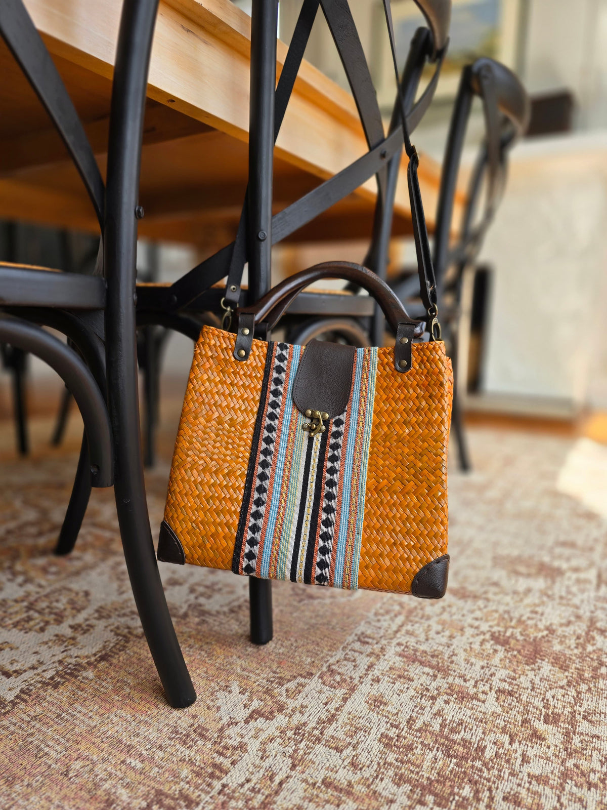 An orange handbag with fabric panel and faux leather tab to fasten it closed. Hanging off a chair around a dining table.