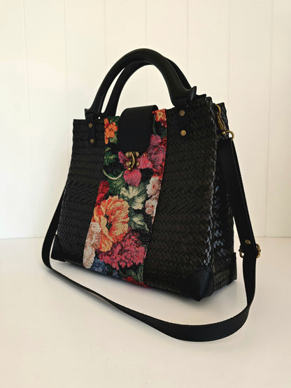 A black woven tote handbag with a vibrant multicolor floral tapestry panel on the front and black wooden handles and faux leather shoulder strap, set against a white paneled background.