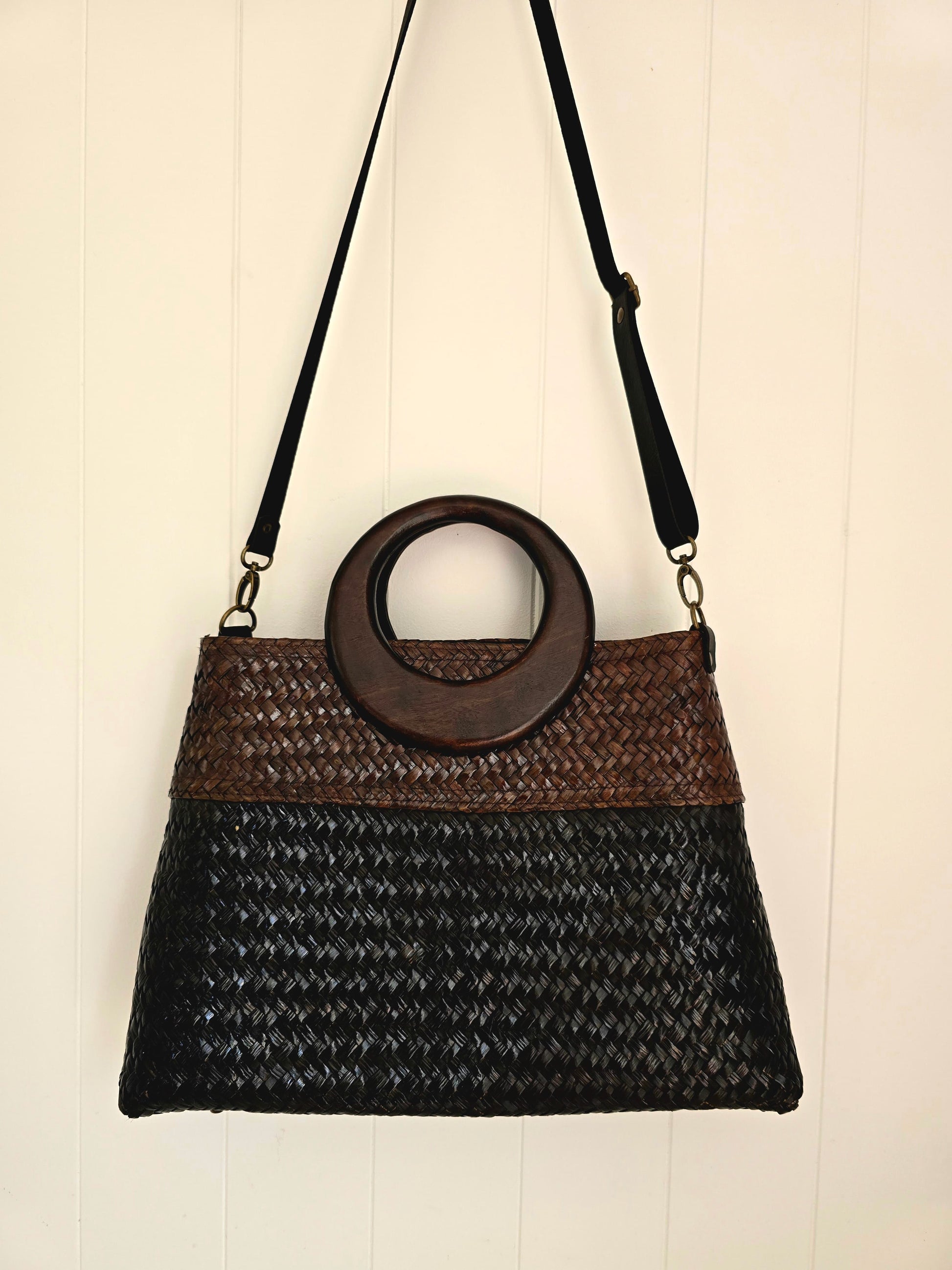 A woven shoulder bag with a black and brown patterned body, a circular dark brown wooden handle, and a black shoulder strap, hanging against a white paneled wall.