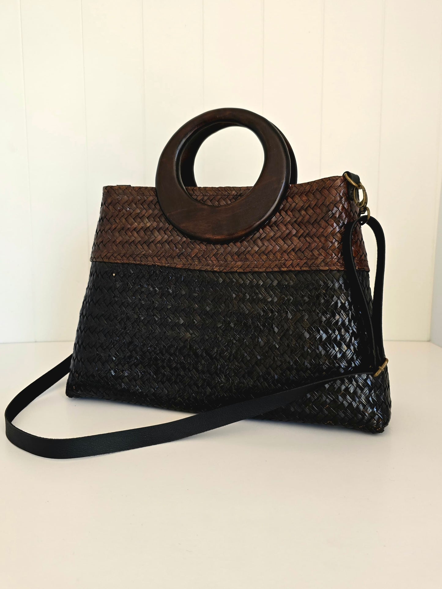 A woven handbag with a two-tone design, featuring a brown woven top section and a black woven bottom section, with a large circular dark wood top handle and a black shoulder strap.