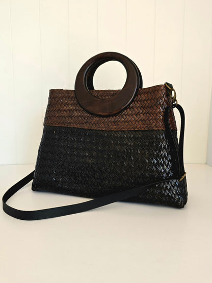A woven handbag with a two-tone design, featuring a brown woven top section and a black woven bottom section, with a large circular dark wood top handle and a black shoulder strap.
