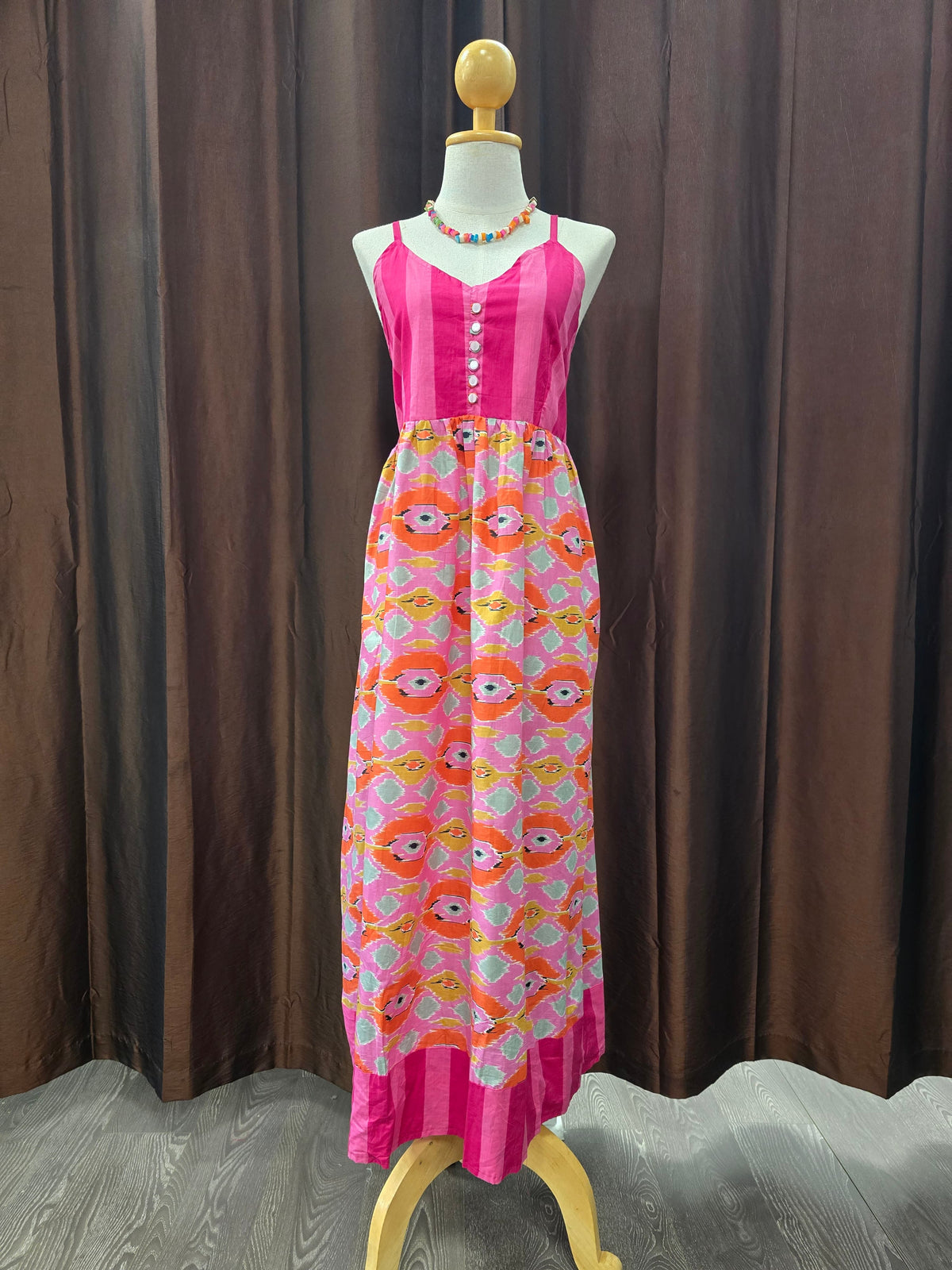 A sleeveless pink maxi dress displayed on a mannequin against a brown curtain backdrop. The dress has a solid bright pink bodice with a button placket and a gathered skirt featuring a repeating cartoon face pattern in pink, orange, yellow, and white.