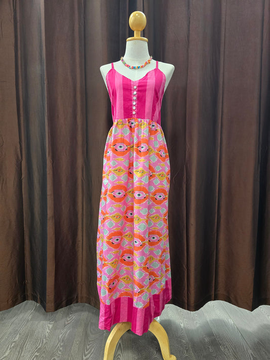 A sleeveless pink maxi dress displayed on a mannequin against a brown curtain backdrop. The dress has a solid bright pink bodice with a button placket and a gathered skirt featuring a repeating cartoon face pattern in pink, orange, yellow, and white.