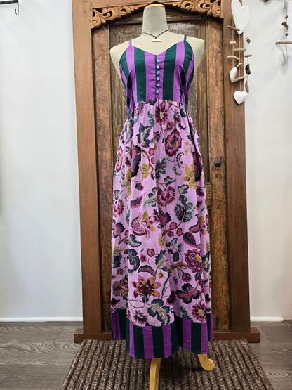 A sleeveless maxi dress displayed on a mannequin in front of a carved wooden door. The dress features a purple and dark green striped bodice with a button front, and a flowing floral skirt in shades of purple, pink, and green.