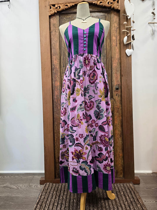 A sleeveless maxi dress displayed on a mannequin in front of a carved wooden door. The dress features a purple and dark green striped bodice with a button front, and a flowing floral skirt in shades of purple, pink, and green.