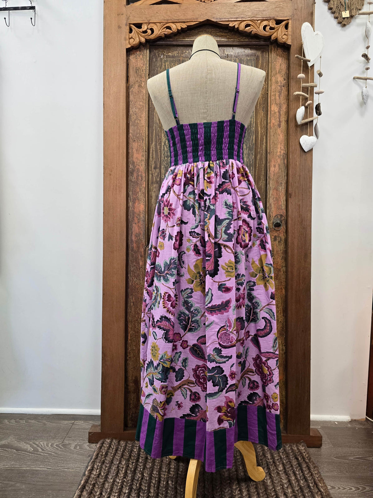 A sleeveless maxi dress displayed on a mannequin in front of a carved wooden door. The dress has a purple and black striped bodice, thin shoulder straps, and a full gathered skirt with a pink, purple, and green floral and butterfly print.