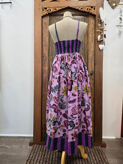 A sleeveless maxi dress displayed on a mannequin in front of a carved wooden door. The dress has a purple and black striped bodice, thin shoulder straps, and a full gathered skirt with a pink, purple, and green floral and butterfly print.