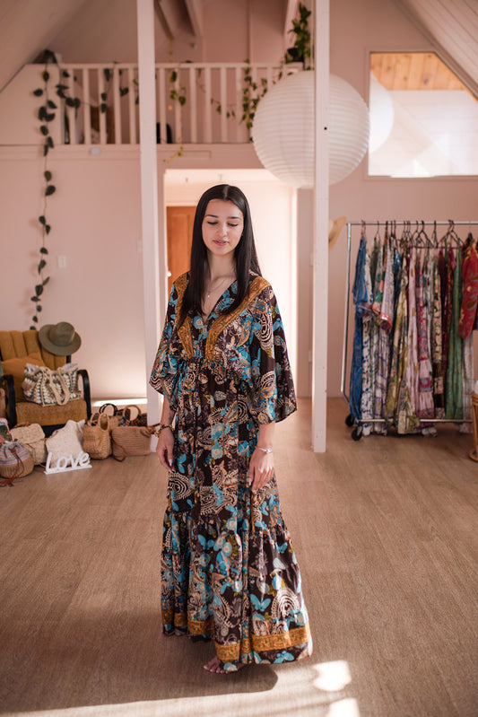 A woman wearing a long bohemian-style maxi dress with a brown, blue, and gold batik print stands in a boutique showroom with wooden floors and clothing racks in the background.