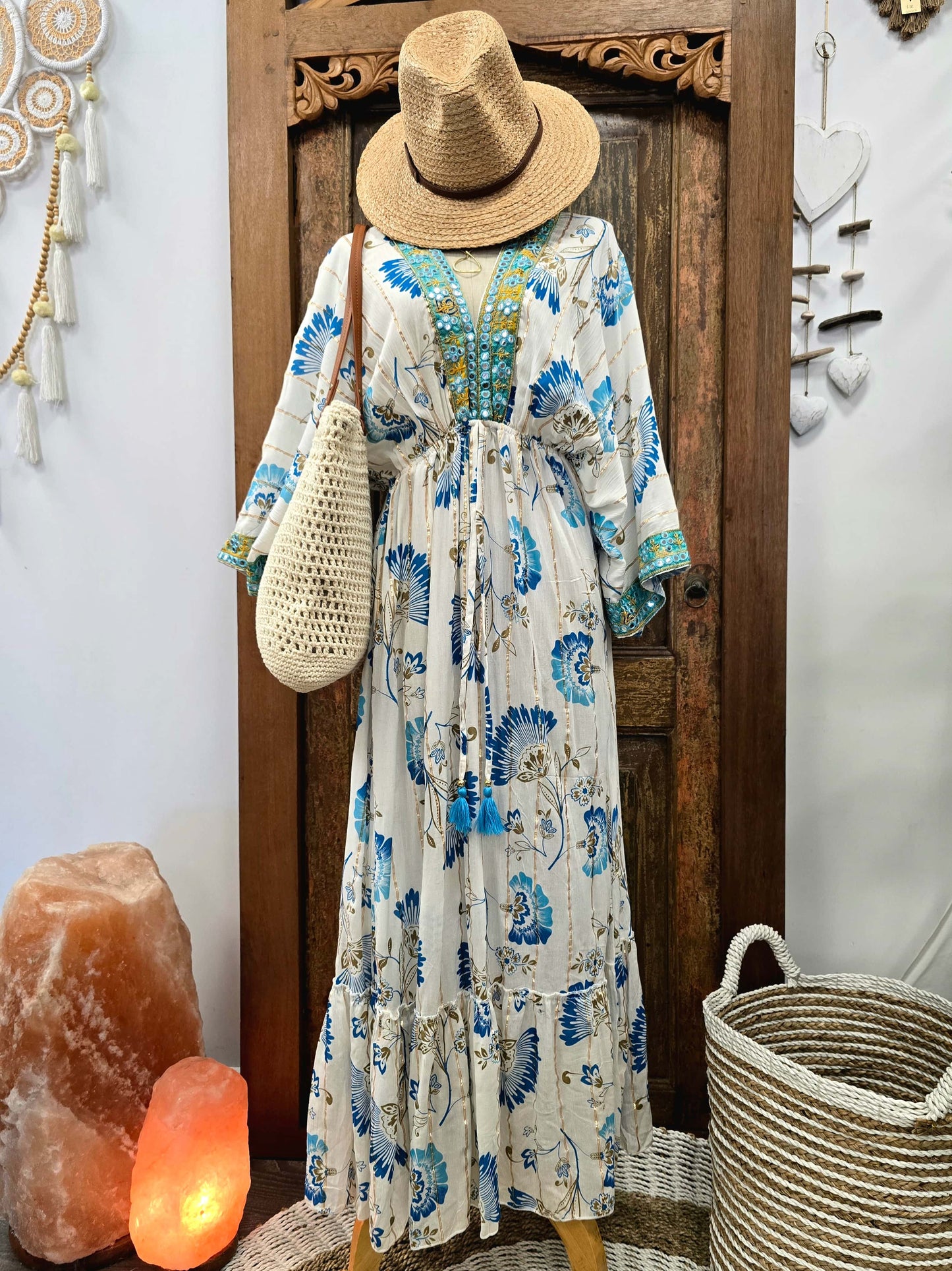 A bohemian style maxi dress with a blue and green floral print on a cream background, styled with a straw hat, woven bag, and accessories in front of a wooden door.