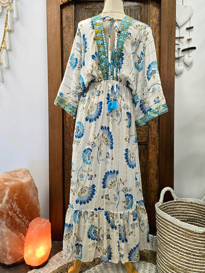 A bohemian-style maxi dress with a blue and green floral print on a cream base, displayed on a mannequin in front of a rustic wooden door, flanked by a salt lamp and a woven basket.