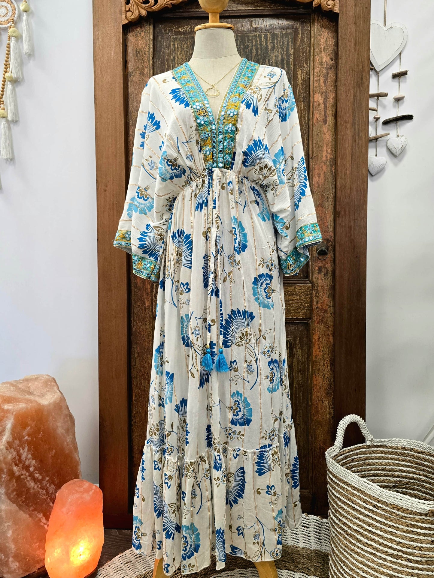 A bohemian style maxi dress with a blue and green floral print on a cream base, displayed on a mannequin in front of a rustic wooden door.