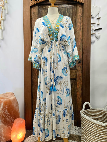 A bohemian style maxi dress with a blue and green floral print on a cream base, displayed on a mannequin in front of a rustic wooden door.