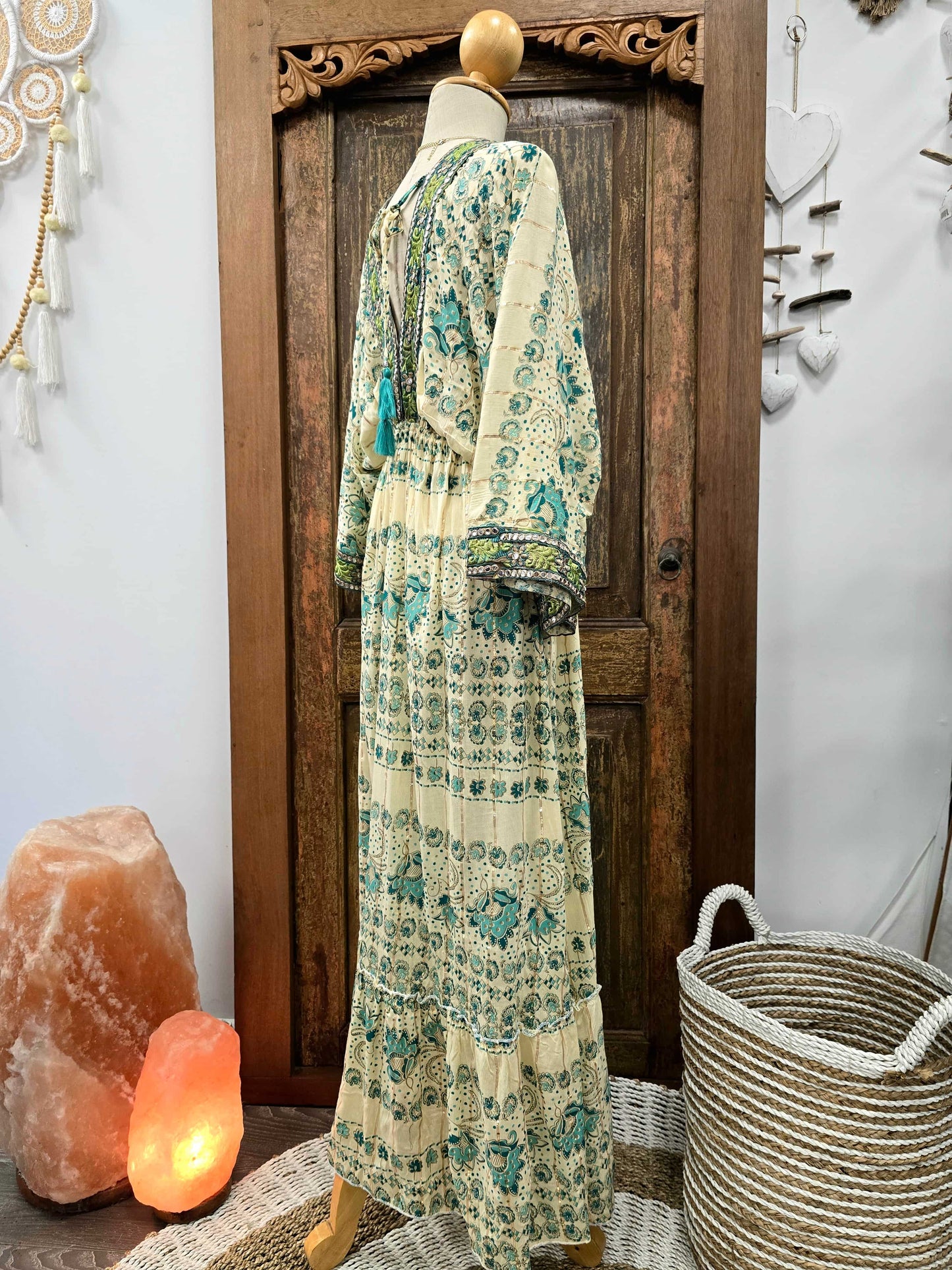 A long cream maxi dress with a teal and green bohemian floral print is displayed on a mannequin in front of a rustic wooden door, with decorative home items like a salt lamp, woven basket, and wall hanging nearby.