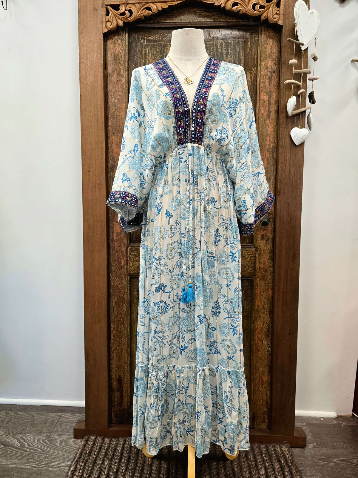 A light blue and white floral print maxi dress with a dark purple embroidered V-neckline, displayed on a mannequin in front of a rustic wooden door.