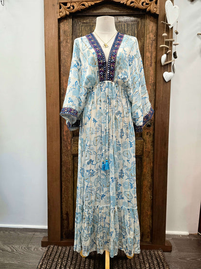 A light blue and white floral print maxi dress with a dark purple embroidered V-neckline, displayed on a mannequin in front of a rustic wooden door.