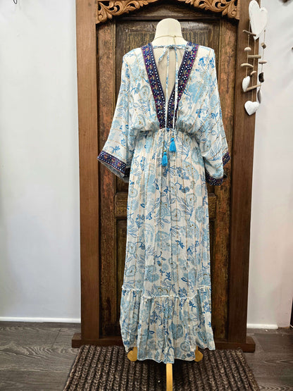 A long kaftan-style dress displayed on a mannequin in front of a carved wooden door. The dress has a light blue and white floral print with dark blue and purple beaded trim along the V-neckline and sleeve cuffs, and blue tassels at the waist.