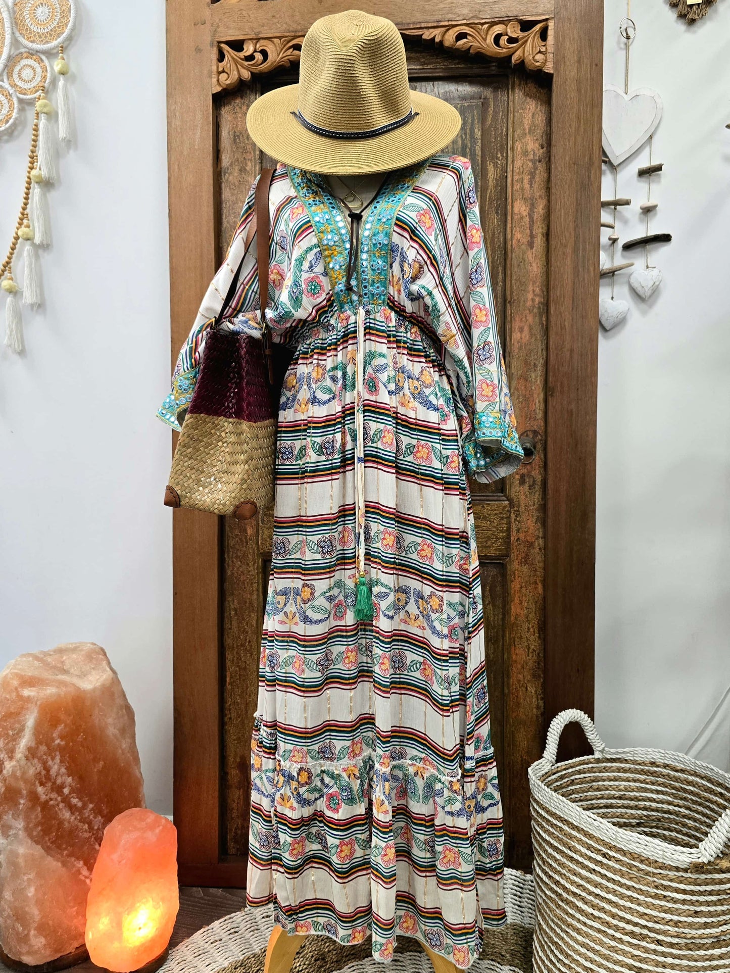 A bohemian-style maxi dress with a colorful paisley and striped pattern, styled on a mannequin with a straw hat and shoulder bag, set against a rustic wooden door background.