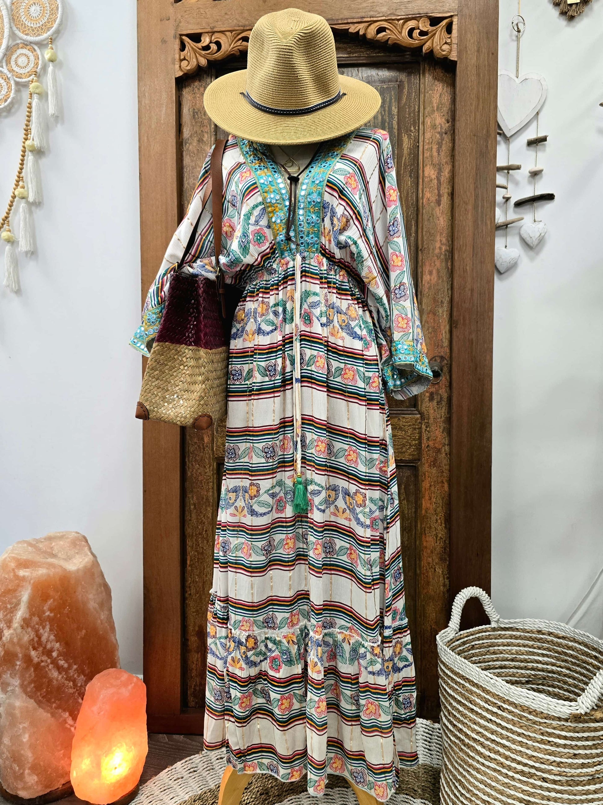 A bohemian-style maxi dress with a colorful paisley and striped pattern, styled on a mannequin with a straw hat and shoulder bag, set against a rustic wooden door background.