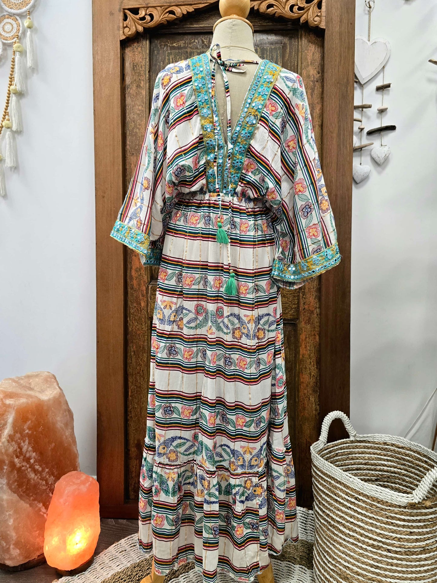 Back view of a bohemian-style maxi dress with a multicolor striped and floral pattern, displayed on a mannequin in front of a wooden door. The dress features a V-neckline with tassel ties and wide sleeves.