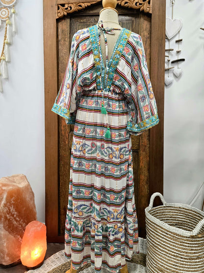 Back view of a bohemian-style maxi dress with a multicolor striped and floral pattern, displayed on a mannequin in front of a wooden door. The dress features a V-neckline with tassel ties and wide sleeves.