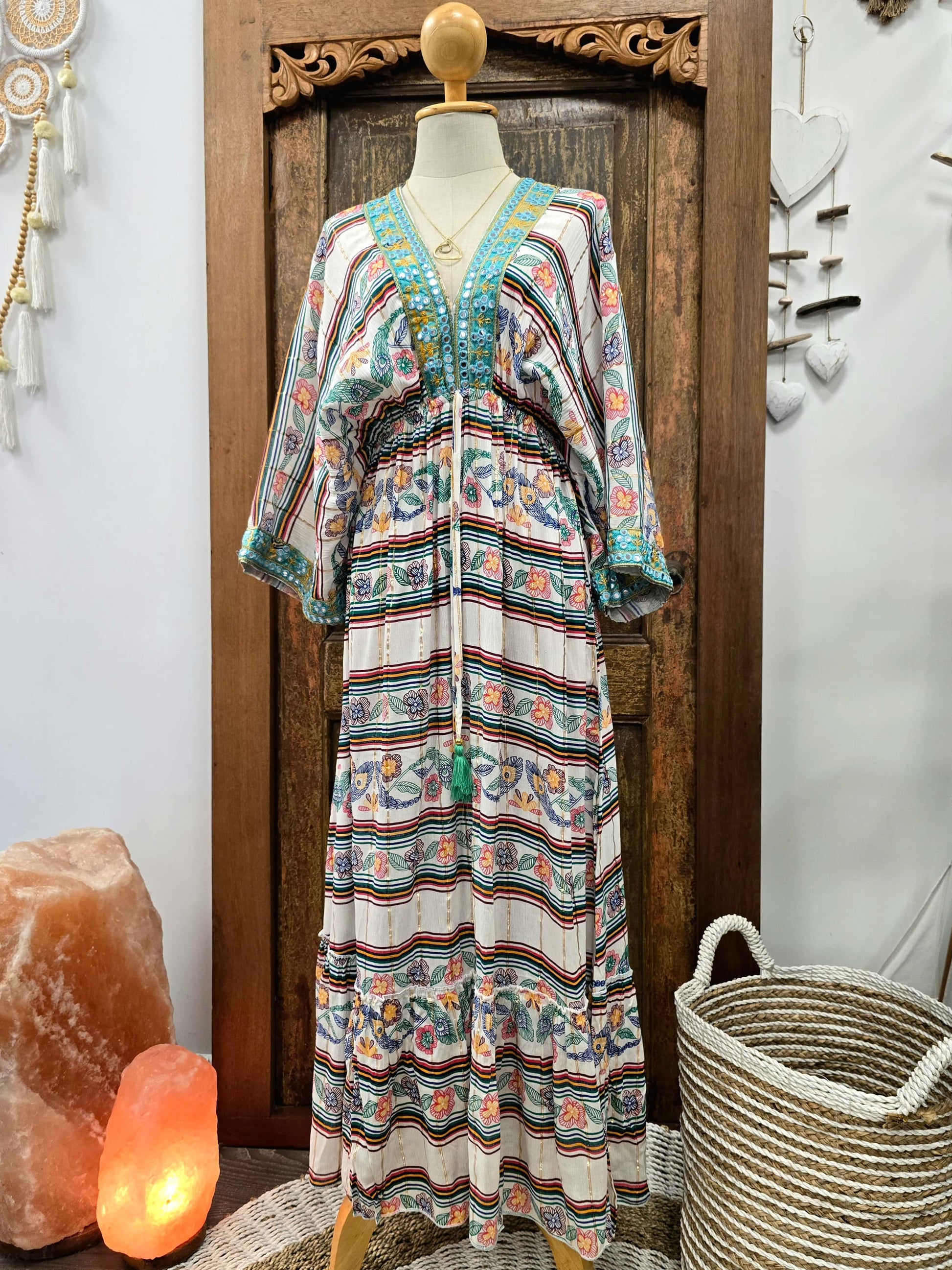 A bohemian-style maxi kaftan dress displayed on a mannequin torso in front of a carved wooden door. The dress features a cream base with horizontal stripes in teal, rust orange, and dark brown, decorated with blue floral motifs and a teal embroidered V-neckline.