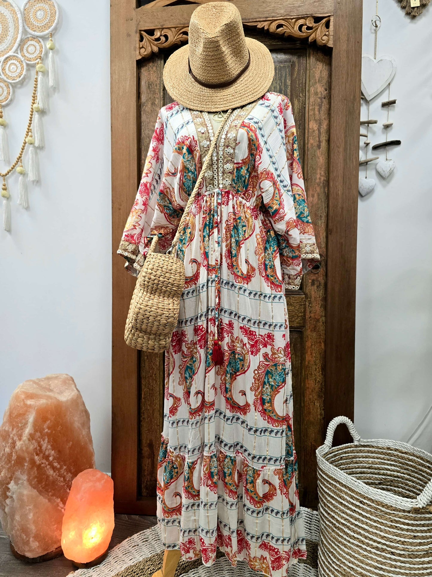 A bohemian-style maxi dress with a red, teal, and blue paisley pattern on a cream background, styled with a straw hat, woven shoulder bag, and straw basket accessories against a rustic wooden door backdrop.