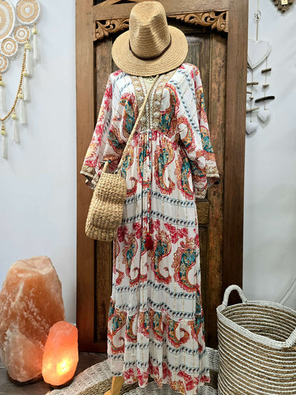 A bohemian-style maxi dress with a red, teal, and blue paisley pattern on a cream background, styled with a straw hat, woven shoulder bag, and straw basket accessories against a rustic wooden door backdrop.