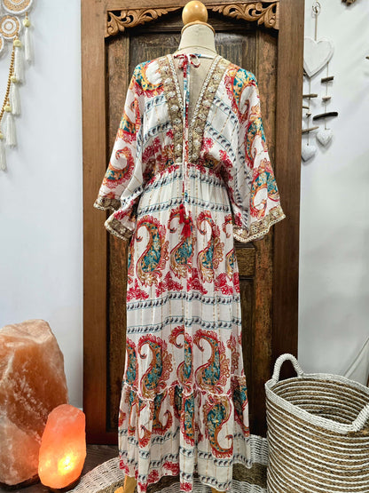 Back view of a bohemian-style maxi dress with a cream base and colorful paisley patterns, displayed on a mannequin in front of a wooden door.