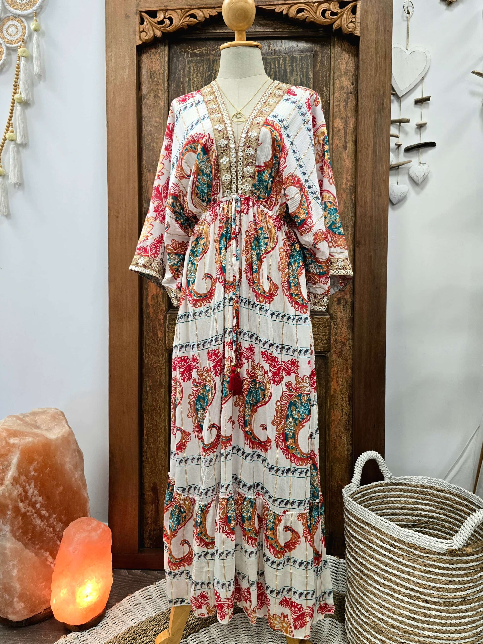 A bohemian-style maxi dress displayed on a mannequin in front of a carved wooden door. The dress features a cream base with a vibrant red, teal, and blue paisley pattern, gold embroidery on the neckline, and wide three-quarter sleeves.
