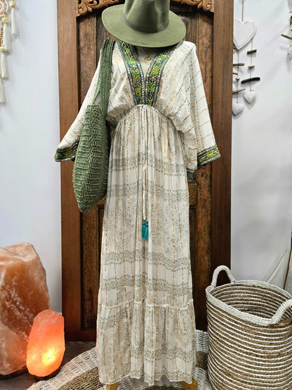 A bohemian-style maxi dress displayed on a mannequin, featuring a cream and beige patterned fabric with green and blue embroidered details around the V-neckline and sleeves. The dress is styled with a matching olive green hat, a green knit shoulder bag, and a long tassel necklace with a turquoise pendant, set against a rustic wooden door background with decorative props like a salt lamp and a woven basket.