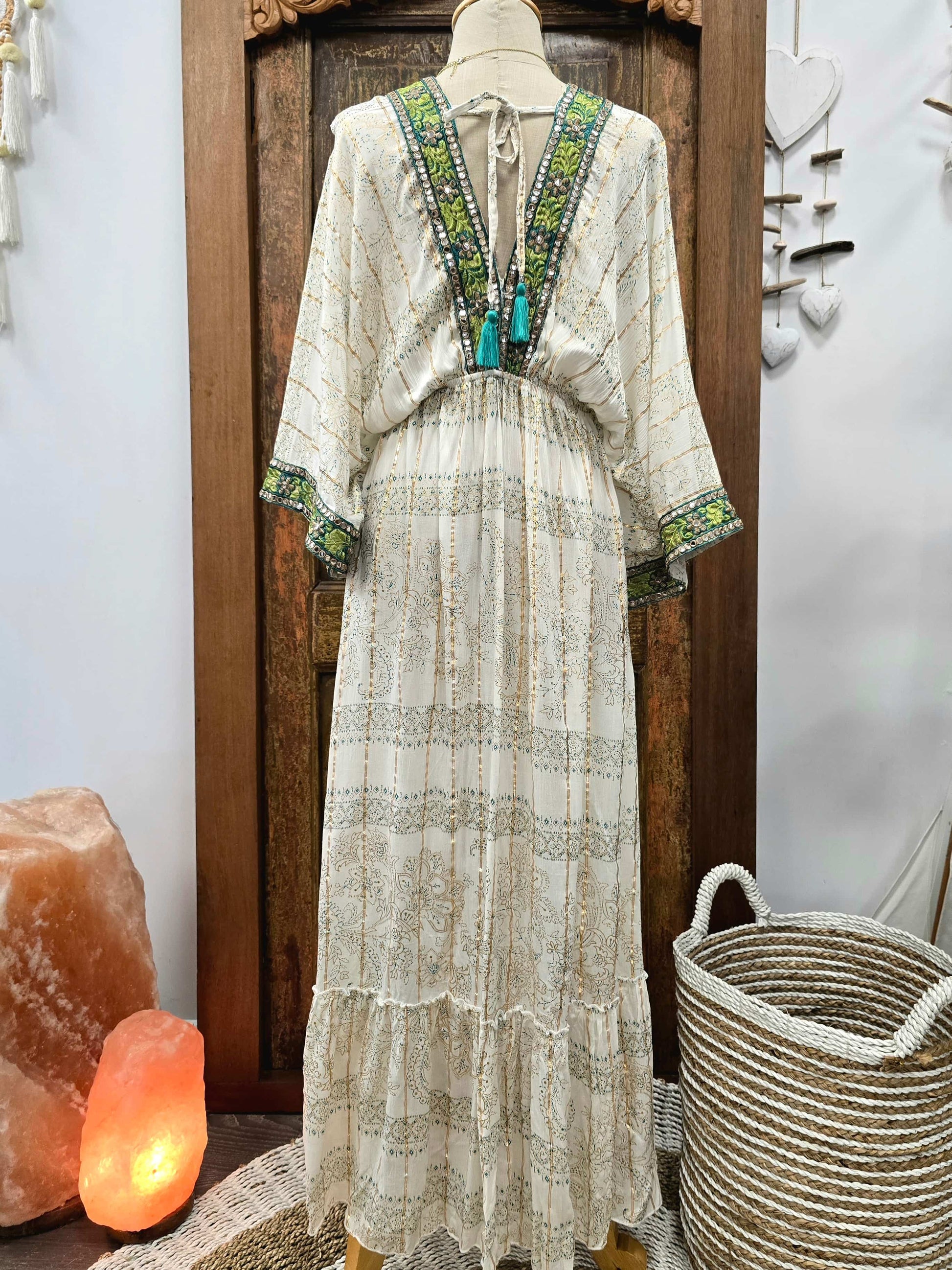 The back of a bohemian-style maxi dress displayed on a mannequin, featuring a cream base with green and turquoise embroidered patterns along the V-neckline and sleeve cuffs. The dress is styled in a rustic indoor setting with a wooden door, a glowing orange salt lamp, and a woven basket.