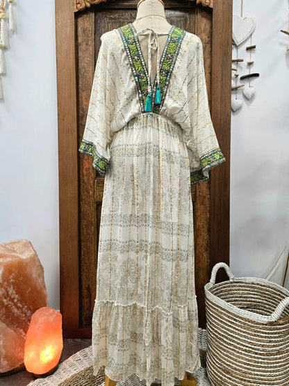 The back of a bohemian-style maxi dress displayed on a mannequin, featuring a cream base with green and turquoise embroidered patterns along the V-neckline and sleeve cuffs. The dress is styled in a rustic indoor setting with a wooden door, a glowing orange salt lamp, and a woven basket.